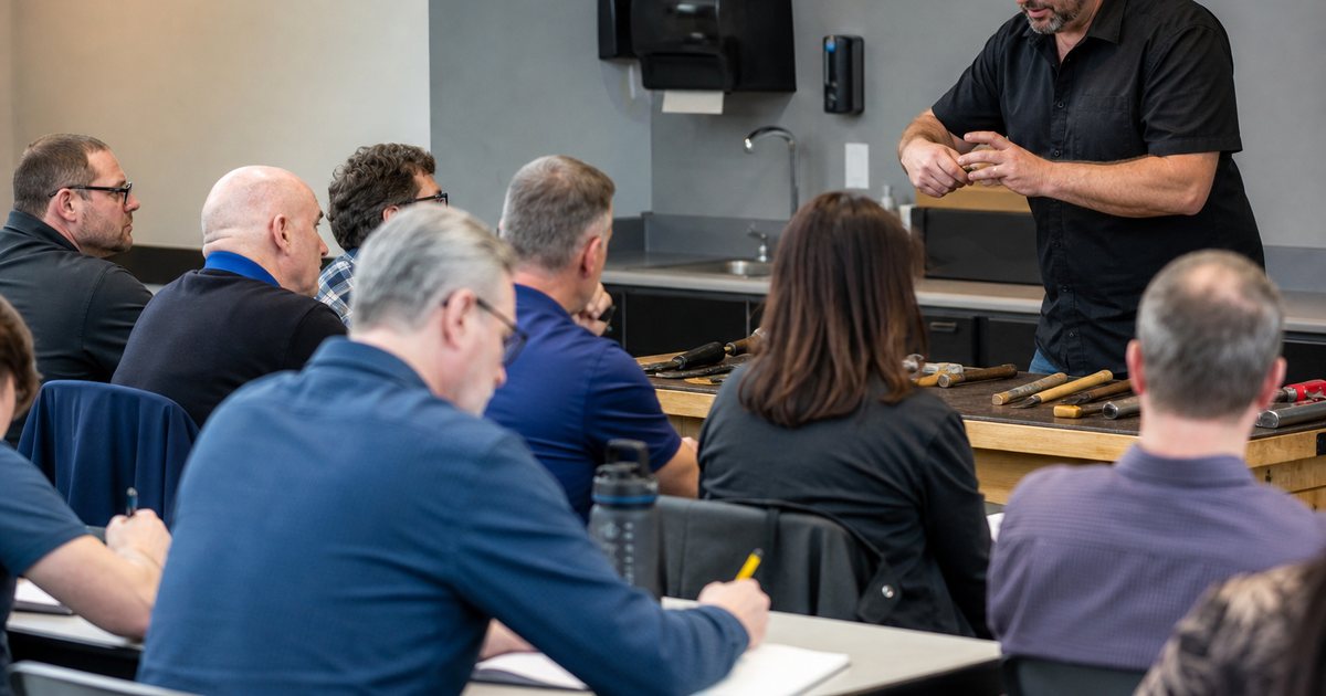 A group of adult professionals in a classroom-style continuing education session, an instructor at the front demonstrating with hand tools on a table, attendees taking notes on paper