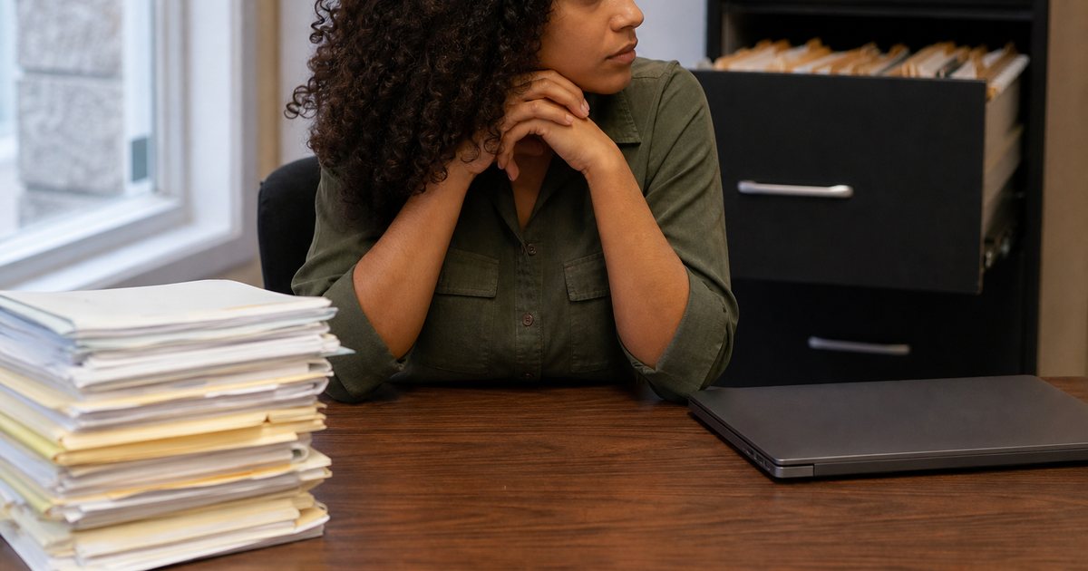 A person at a desk in a small office with stacks of paper personnel folders on one side and a closed laptop on the other, a filing cabinet with drawers open in the background