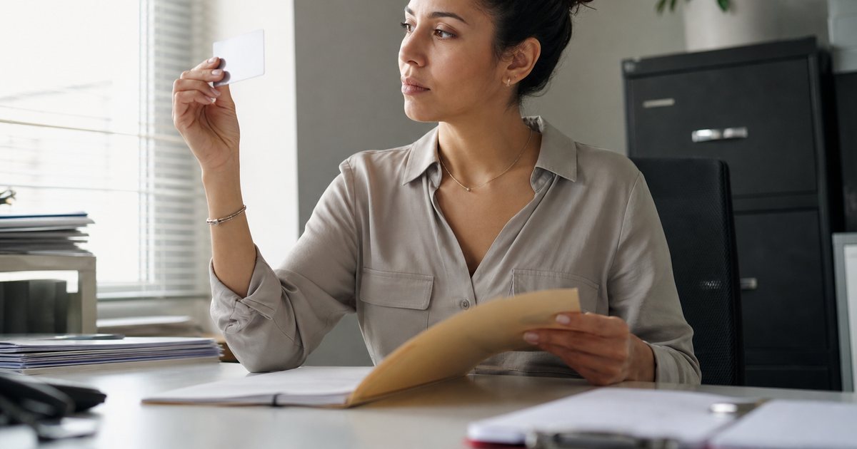 An HR coordinator at a desk in a small office on a desk phone call, a new employee seated across from her waiting, a closed folder between them, office plants and a filing cabinet in the background